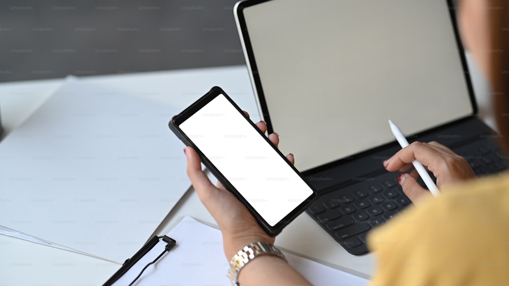 Immagine ritagliata di una bella donna che lavora come segretaria con in mano uno smartphone con schermo bianco e una penna stilo mentre è seduta davanti al suo tablet con custodia per tastiera sopra l'ufficio come sfondo.