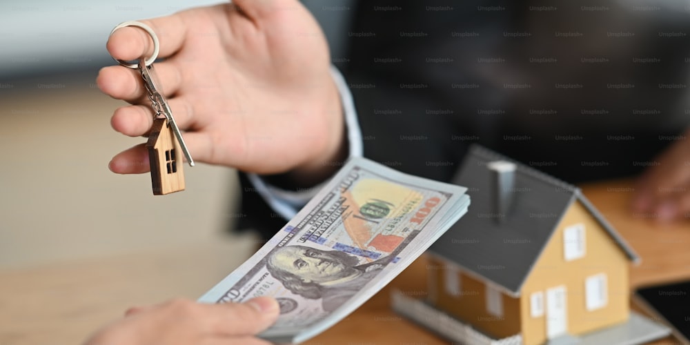 Cropped image of Real estate agent offer house insurance or house ownership to his customer at the modern wooden table. Hands giving a houses key and banknote. Trading/Dealing concept.