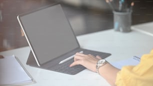 Cropped image of creative woman holding a stylus pen while drawing/pointing on white blank screen computer tablet with keyboard case that putting on working desk over comfortable room as background.