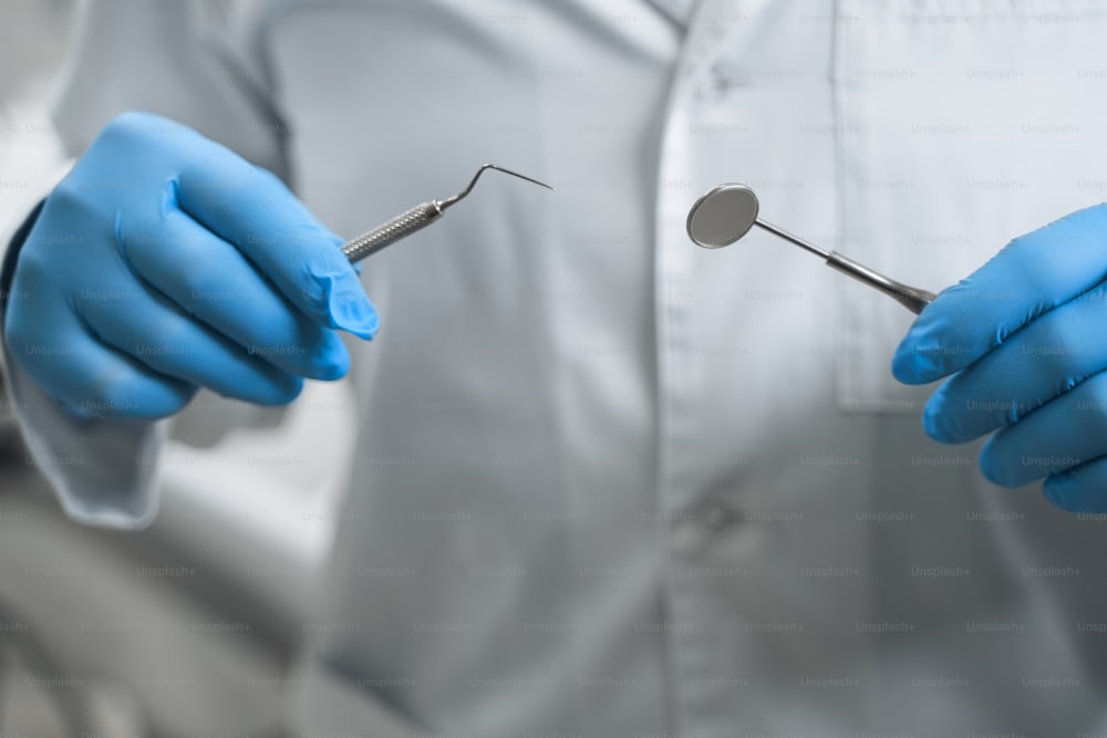 Male specialist in white uniform is holding dental explorer and mirror while wearing sterile gloves