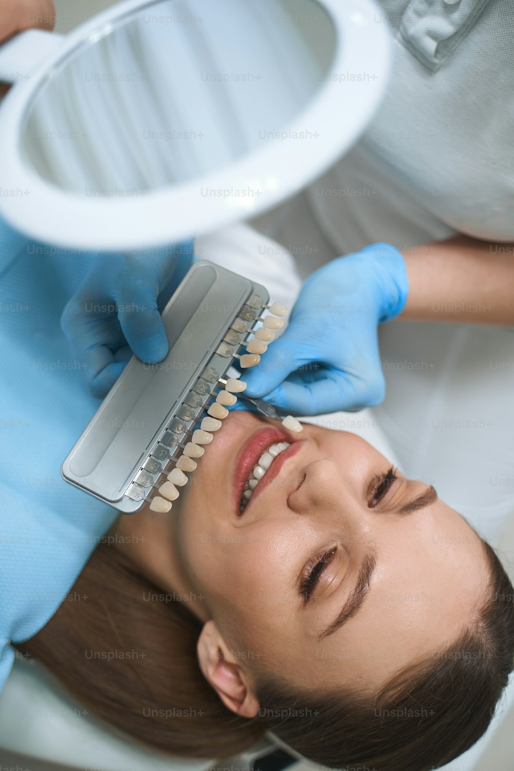 Smiling young woman is lying in chair in dental clinic and choosing veneers with doctor assistance