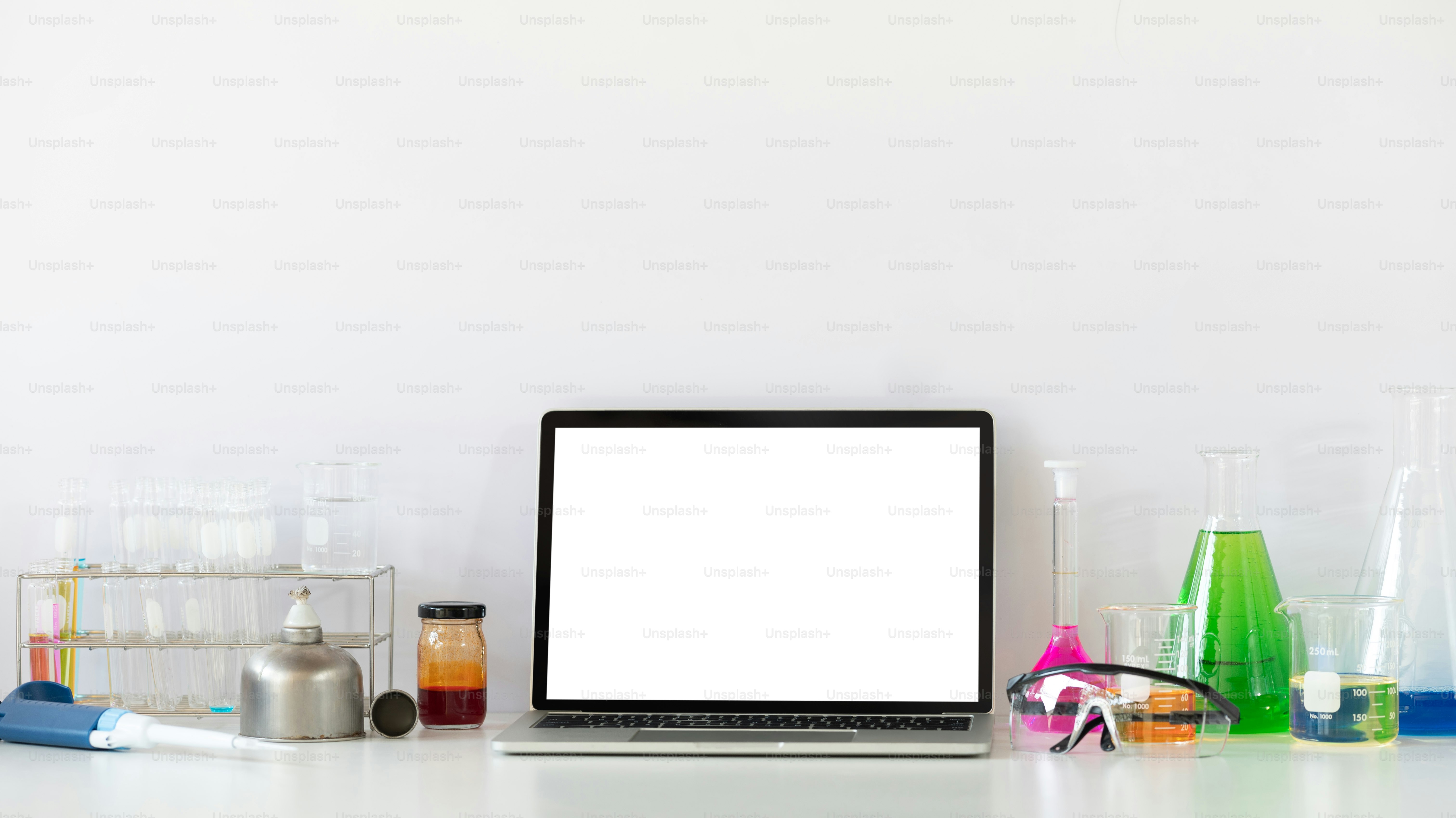Photo of Scientific experiments equipment putting on white working desk with white blank screen computer laptop. Flat lay computer laptop, chemistry glassware, safety glasses.