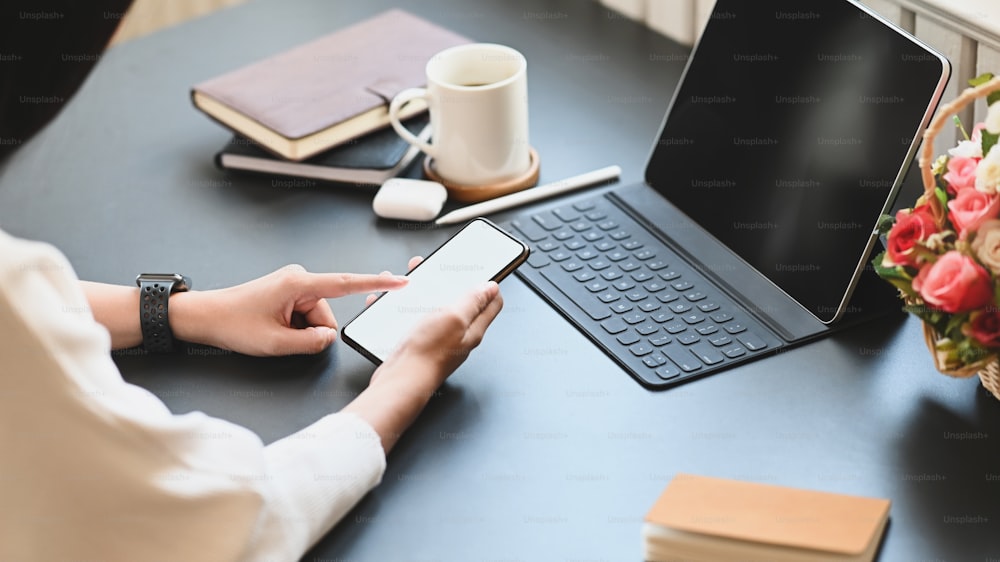 Cropped image of beautiful woman working as secretary holding/using a white blank screen smartphone while sitting in front her computer tablet with keyboard case over office as background.