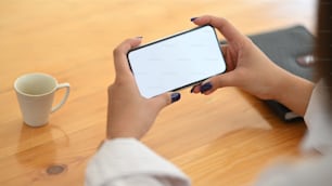 Imagen recortada de las manos de una joven creativa sosteniendo un teléfono inteligente con la pantalla vacía mientras está sentada en el escritorio de trabajo sobre una cómoda sala de estar como fondo.