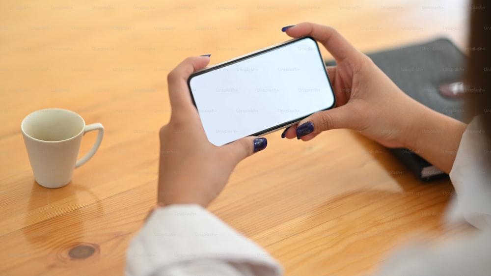 Immagine ritagliata delle mani di una giovane donna creativa che tiene uno smartphone con schermo vuoto mentre è seduta alla scrivania sopra un comodo soggiorno come sfondo.