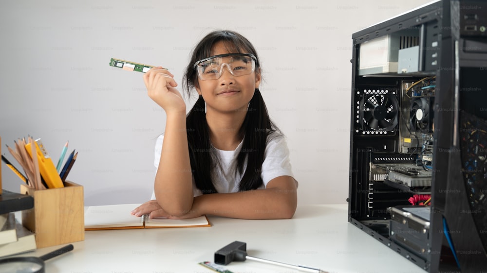 Adorable écolière apprenant à réparer un matériel informatique qui met sur un bureau de travail blanc avec un tournevis et un équipement de technicien sur un mur isolé blanc comme arrière-plan.