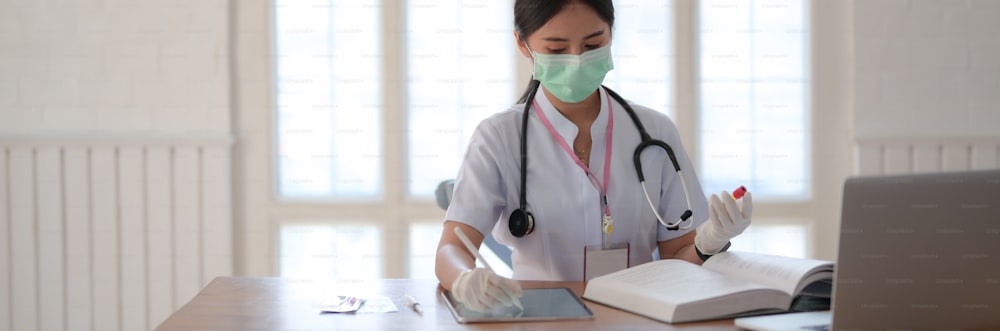 Plan recadré d’une femme médecin tenant un tube à essai et recherchant des informations dans un livre médical et un ordinateur portable tout en écrivant sur une tablette numérique