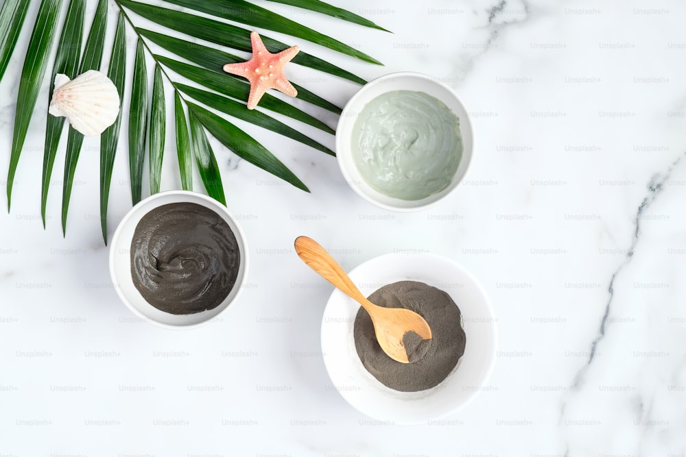 Facial clay mask and powder in bowls with tropical palm leaf on marble background. SPA natural organic cosmetic products for face skin care and treatment. Flat lay, top view.