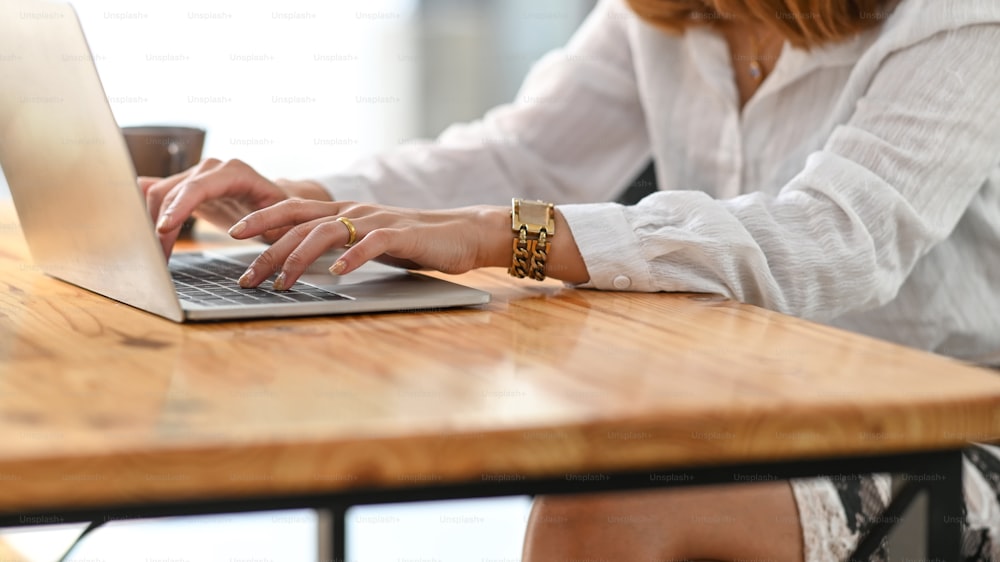 Imagen recortada de una hermosa mujer que trabaja como secretaria escribiendo en una computadora portátil mientras está sentada en el escritorio de trabajo de madera sobre un lugar de trabajo cómodo como fondo.