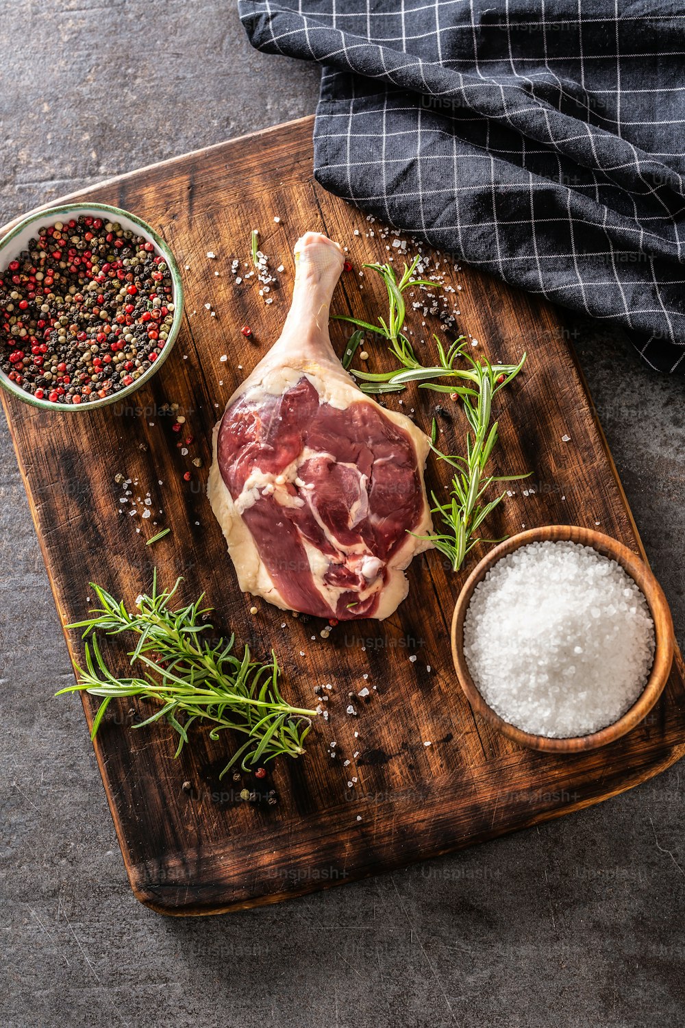 Vue de dessus en format vertical d’une cuisse de canard sur une planche de bois et fond métallique, entourée de nappe et d’ingrédients d’assaisonnement.