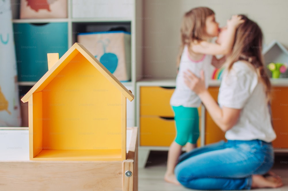 Stay home with family concept. Yellow wooden toy house with copy space on the table in the kids room. Defocused toddler daughter kissing mother.