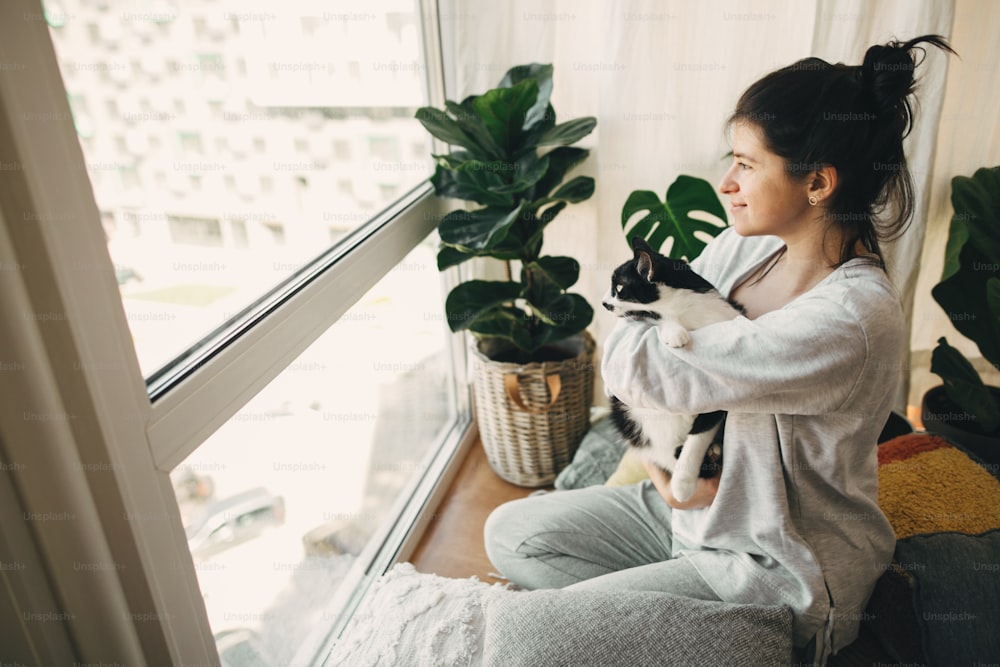 Quédate en casa, mantente a salvo. Chica hipster abrazando a un lindo gato, sentados juntos en casa durante la cuarentena por coronavirus.  Aislamiento en casa para prevenir la epidemia del virus. Mujer joven con gato en habitación moderna