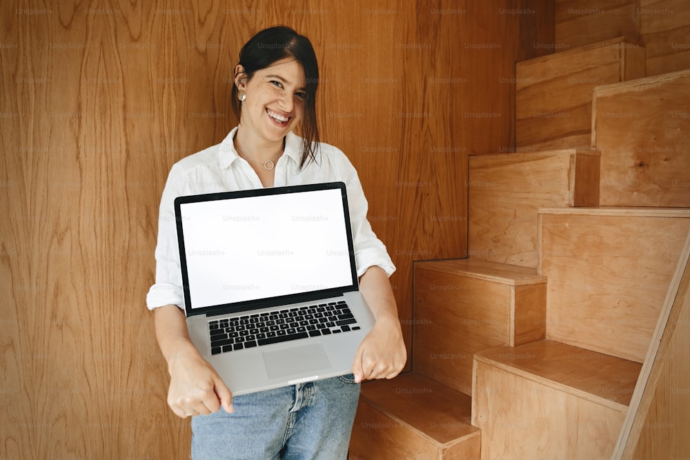 Happy stylish girl showing laptop with empty screen, standing at wooden stairs in modern room. Young business woman working online and smiling. Mockup. Marketing and presentation