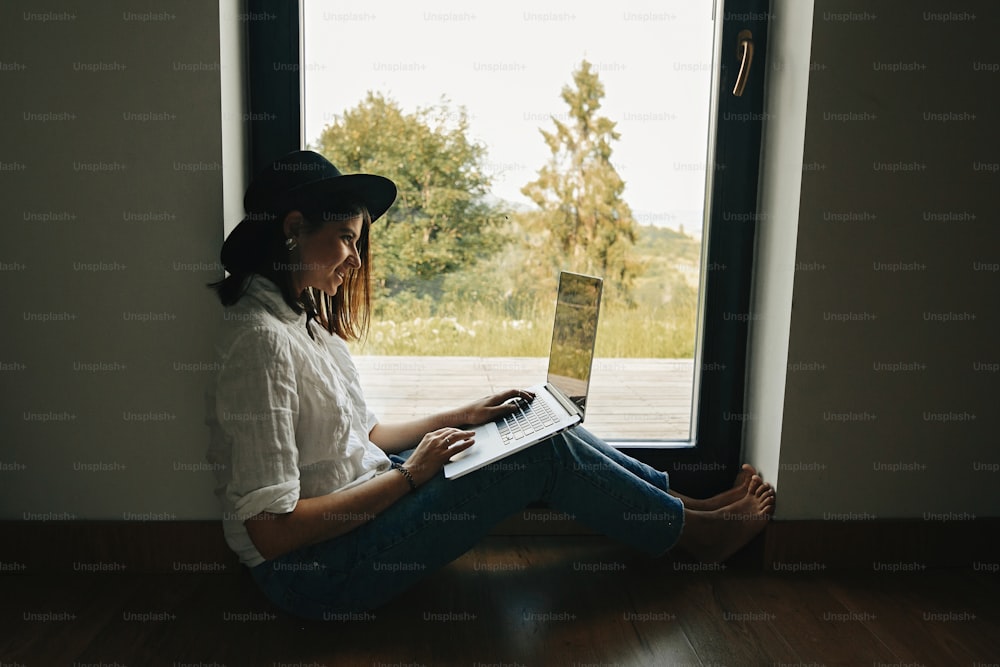 Stylish hipster girl with laptop sitting on floor at big window with view into woods. Young happy woman in hat using laptop, shopping or working online from home. Freelance and freelancer