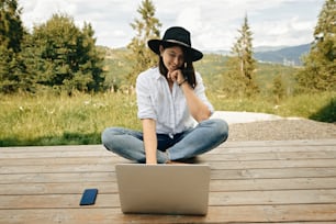 Travel and Freelance. Hipster girl using laptop, sitting on wooden porch with beautiful view on woods and mountains. Stylish woman in hat blogging, shopping or working online outside.
