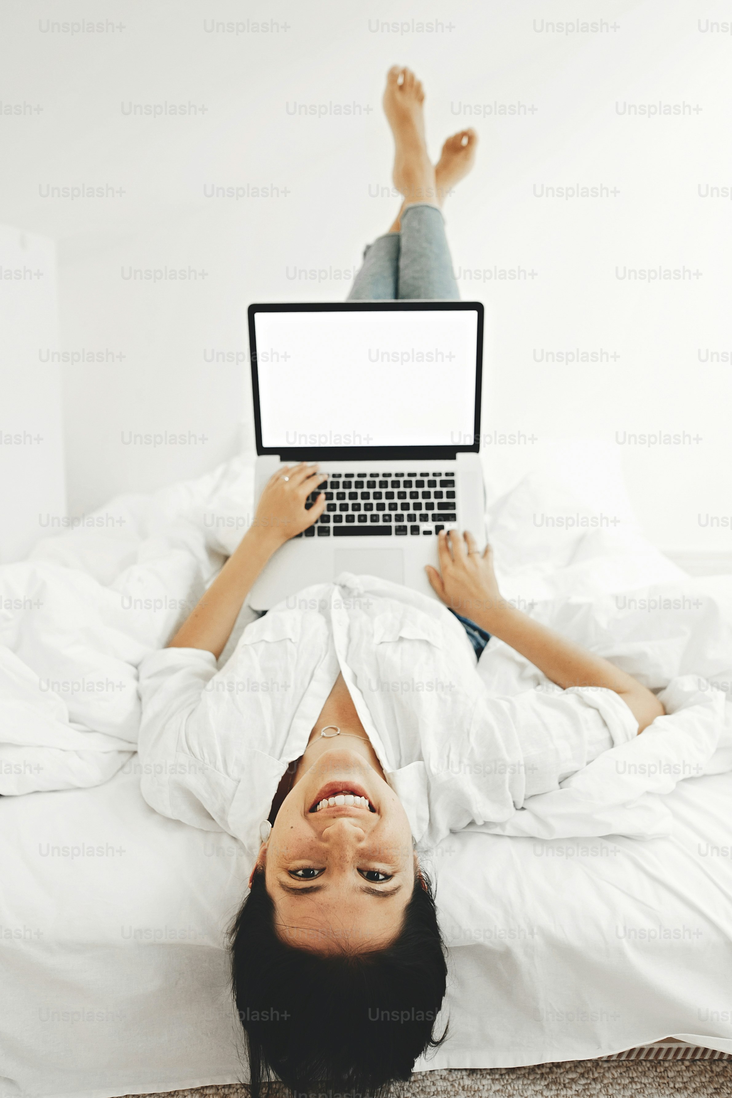 Happy stylish girl smiling, lying on bed with laptop with blank screen in white modern room. Young cheerful woman shopping online or working online from home and smiling. Freelance mockup
