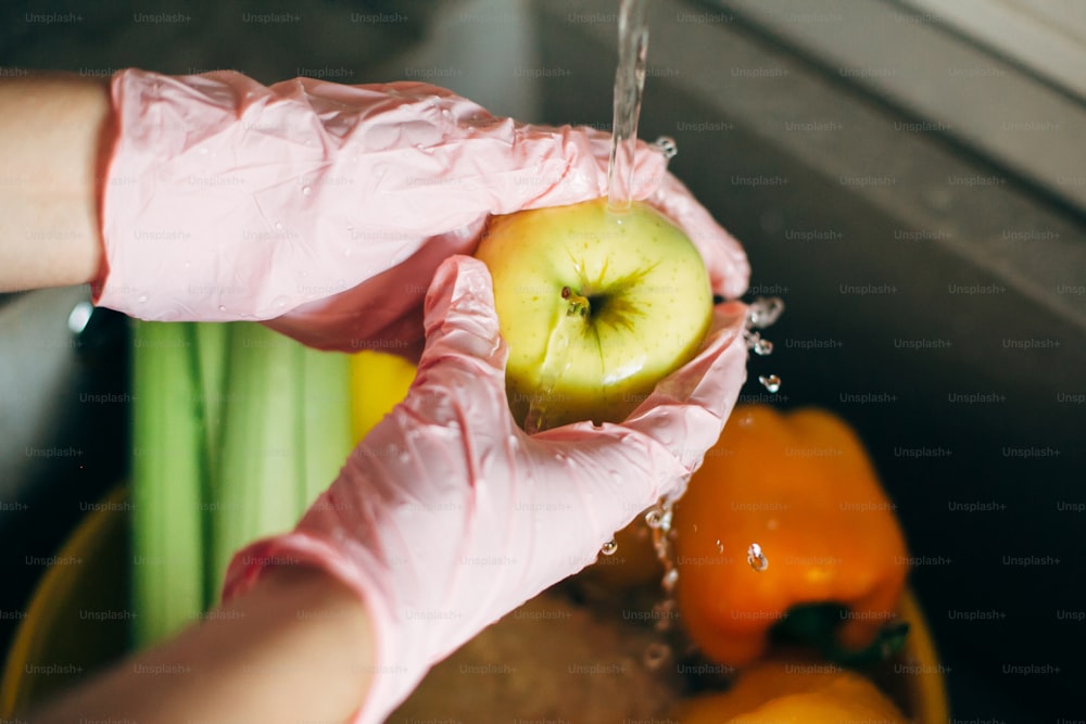 Lavar frutas. Mãos em luvas cor-de-rosa lavando maçã na corrente de água na pia durante a epidemia de vírus. Mulher que limpa legumes e frutas frescos, preparando para cozinhar a refeição na cozinha moderna