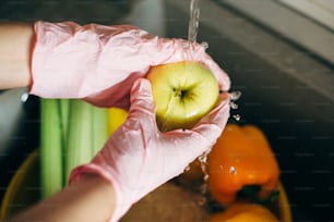 과일 씻기. 분홍색 장갑을 낀 손은 바이러스 전염병 동안 싱크대의 물줄기에서 사과를 씻습니다. 신선한 야채와 과일을 청소하고 현대적인 주방에서 식사를 준비하는 여자