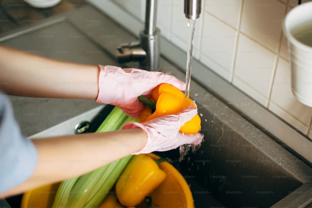Washing vegetables. Hands in gloves washing pepper under water stream in sink during virus epidemic. Woman in pink hands cleaning fresh vegetables, preparing for cooking meal in modern kitchen