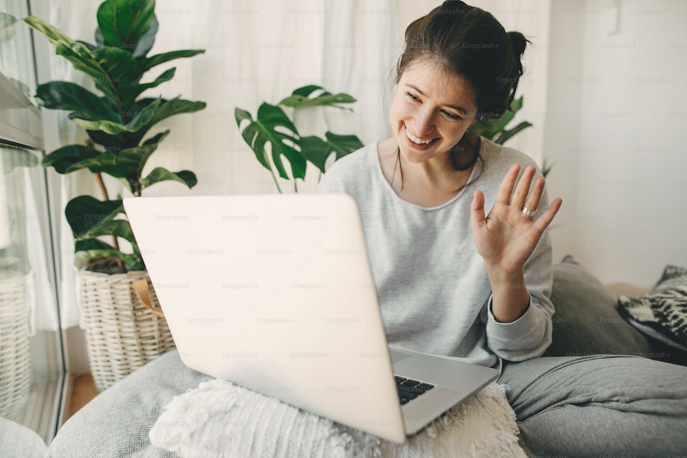 Happy girl waving hi to laptop, video chatting with family or friends, sitting in modern room with pillows and plants. Young casual woman using laptop for communication in quarantine. Home office