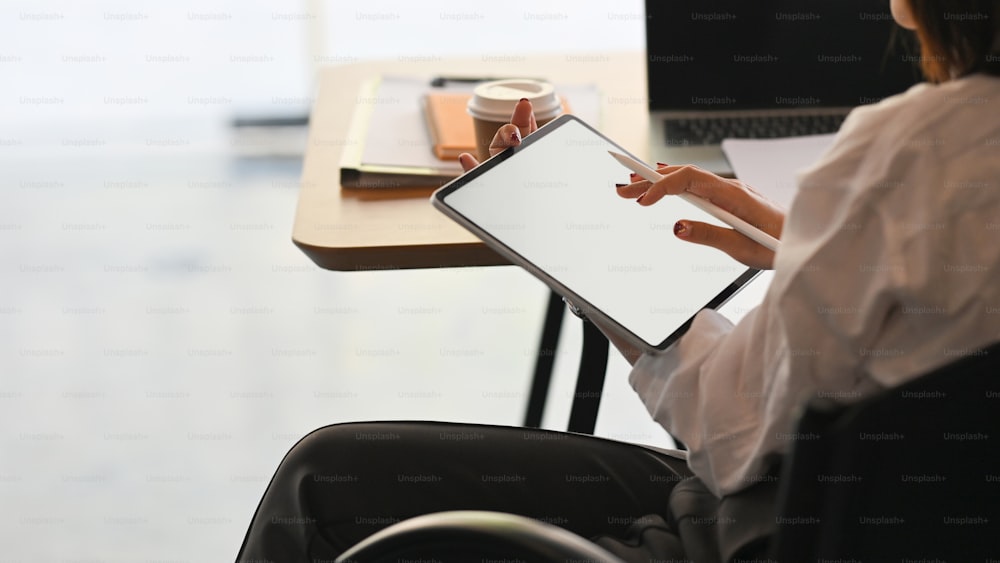 Cropped image waist up of secretary woman holding a stylus pen while sitting and using a white blank screen computer tablet over the modern working table as background.
