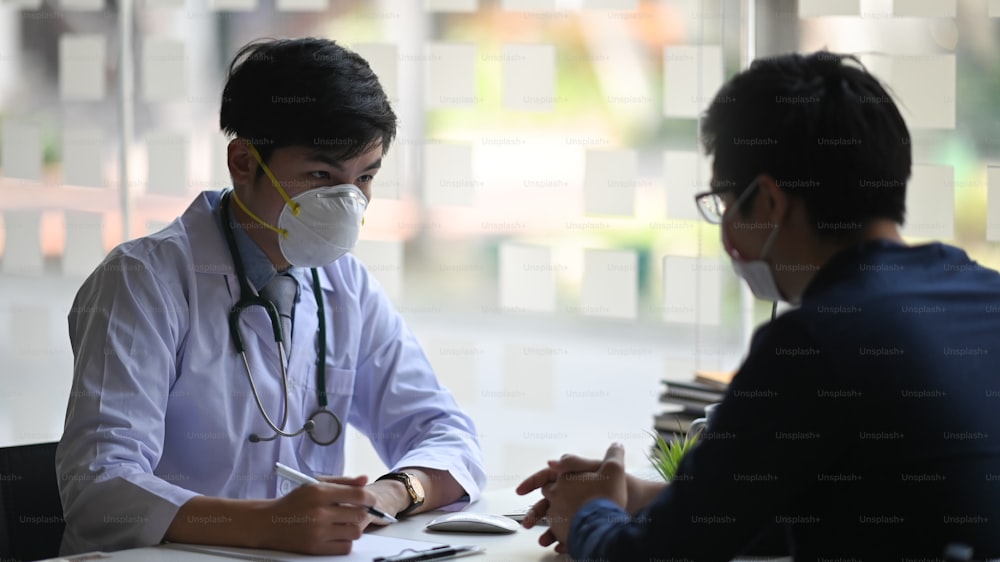 Photo of young smart doctor giving an advice to his patient while sitting together at the doctor working desk over orderly examination room as background. Diagnosis/Disease examination concept.