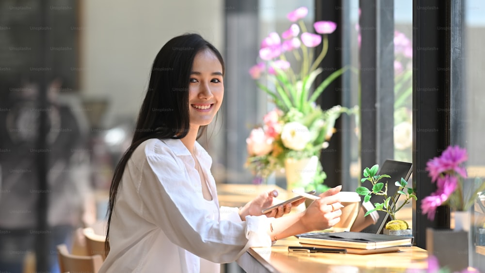 Photo of gorgeous woman in white shirt working with stylus pen and computer tablet while sitting at the wooden counter bar over modern cafe/restaurant as background.