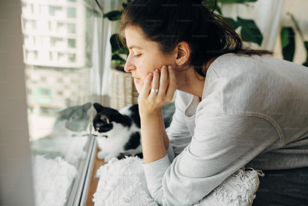 コロナウイルスの検疫中に家に座っている猫と一緒に窓を見ている若い女性。家にいて、安全を確保してください。ウイルスの流行を防ぐための自宅での隔離。モダンな部屋に猫とヒップスターの女の子