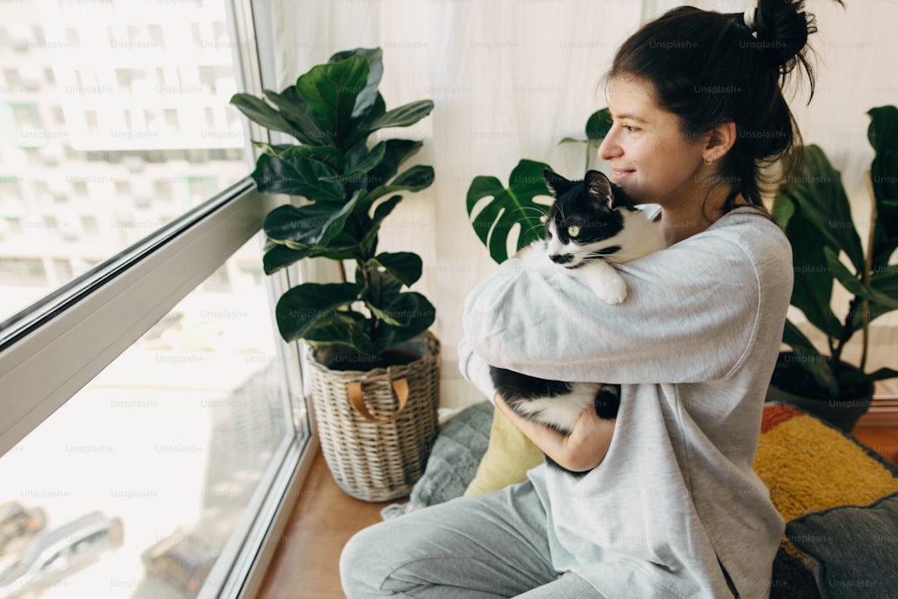 Happy hipster girl hugging cute cat, sitting together at home during coronavirus quarantine. Stay home stay safe. Isolation at home to prevent virus epidemic. Young woman with cat in modern room