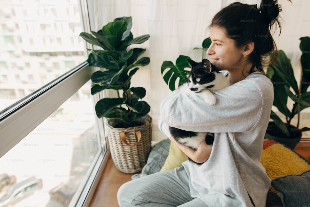 Glückliches Hipster-Mädchen, das eine süße Katze umarmt und während der Coronavirus-Quarantäne zu Hause sitzt. Bleiben Sie zu Hause und bleiben Sie sicher. Isolation zu Hause, um eine Virusepidemie zu verhindern. Junge Frau mit Katze in modernem Zimmer