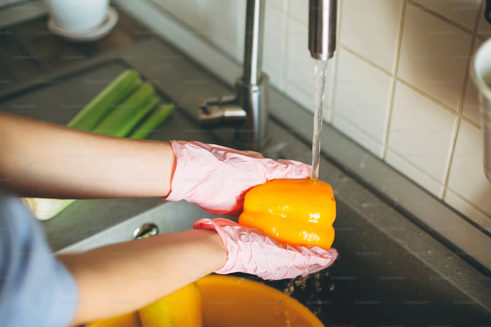 Mãos em luvas segurando pimenta, lavando legumes durante a epidemia de vírus. Mulher nas mãos cor-de-rosa lavando legumes frescos, preparando para cozinhar a refeição na cozinha moderna
