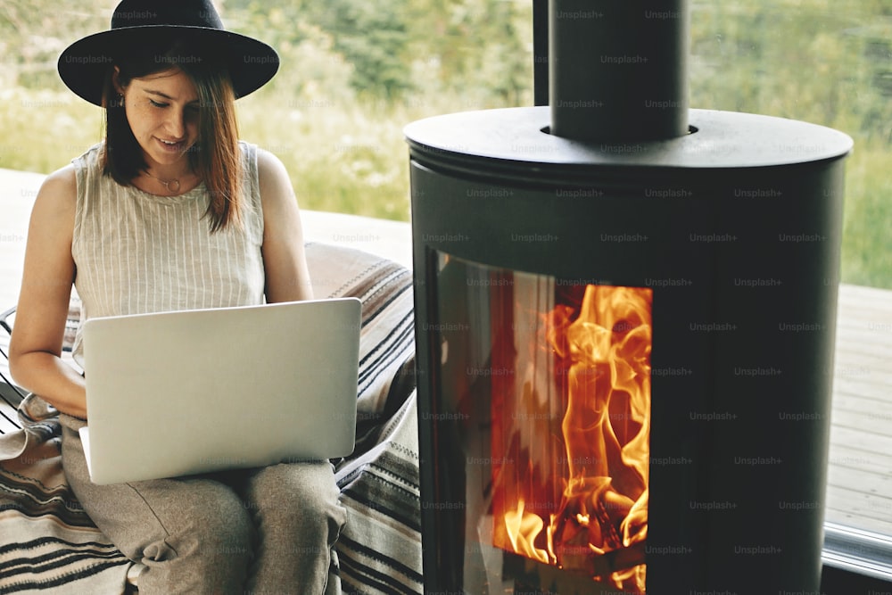 Stylish hipster girl sitting with laptop on cozy chair near fireplace with fire in modern living room with big windows. Working home online. Freelance and freelancer concept. Leisure time