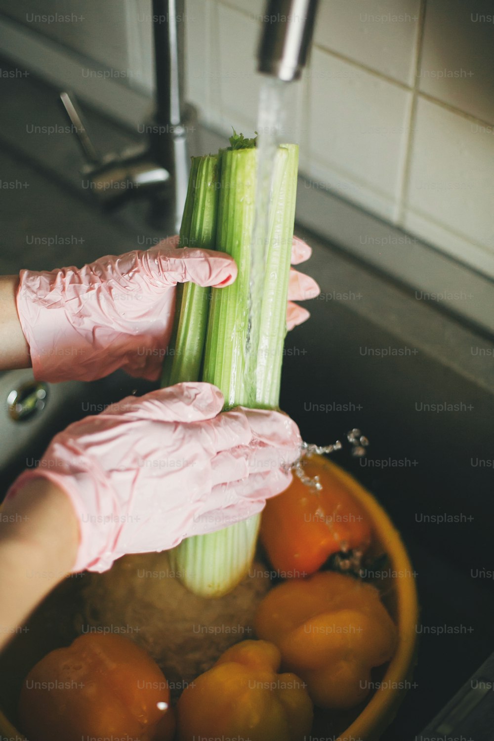 Lavar verduras. Manos con guantes rosas lavando apio en un arroyo de agua en el fregadero durante la epidemia de virus. Mujer limpiando verduras frescas, preparándose para cocinar la comida en la cocina moderna