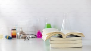 과학 장비, 화학 유리 제품 및 책 더미가 실험실 흰색 벽 위의 흰색 작업 책상에 배경으로 모여 있습니다.