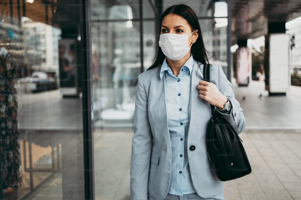Young and elegant business woman looking at storefront on empty city street while wearing protective mask to protect herself from dangerous flu or virus. Corona virus or Covid-19 concept.