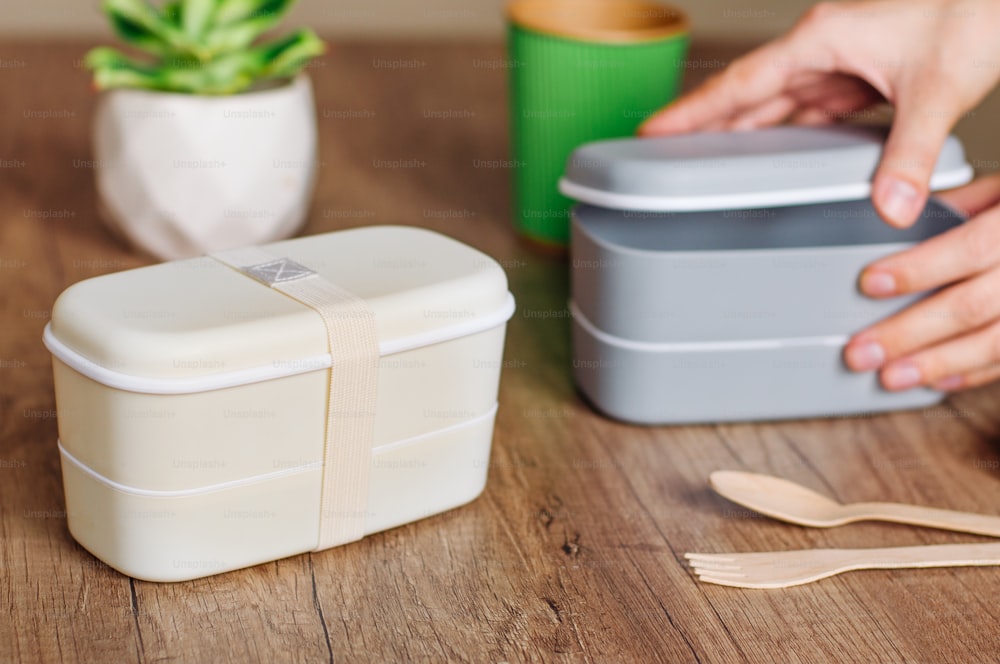 Two closed plastic two-layered lunch boxes with elastic band near wooden forks and spoons and paper cup on wooden table. Female hands opening lunch box on the background. Selective focus on lunch box on the table.