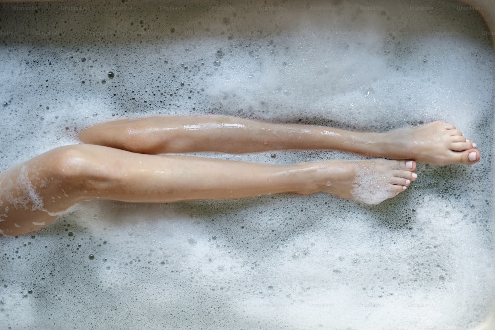 바디 케어와 스킨 케어 개념. 거품 물 위에 다리를 잡고 집에서 목욕을 하는 젊은 성인 소녀의 높은 각도와 잘린 보기