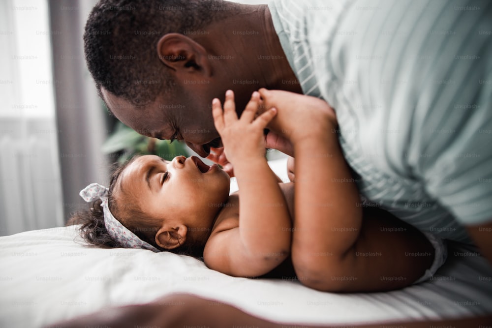 사랑스러운 아프리카계 미국인 아기 소녀가 침대에 누워 있는 동안 아빠는 그녀를 보고 웃고 스톡 사진