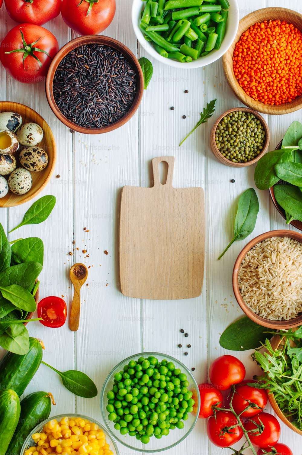 Healthy food concept. Wooden cutting board surrounded by fresh vegetables, groats, leafy greens and quail eggs on white wooden concept with blank space for text. Top view, flat lay.