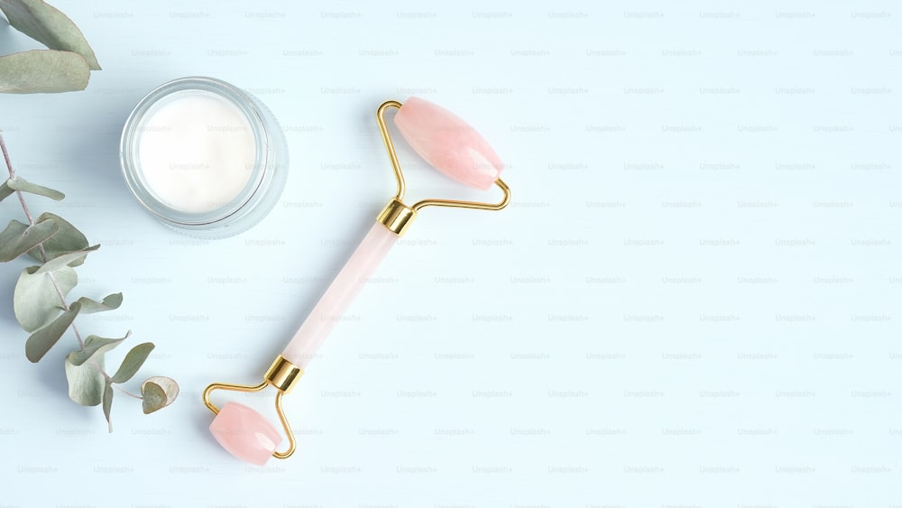 Rose quartz facial massage roller with moisturizer cream and eucalyptus leaf on blue background. Face massager with jade stone, anti-aging, anti-wrinkle beauty skincare tool