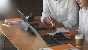Cropped image of business developer team discussing/planning/meeting by using a computer tablet and laptop while sitting together at the wooden meeting table over modern office as background.