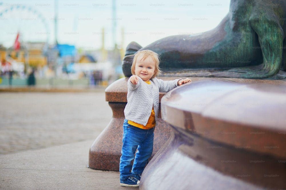 Little baby girl standing close to city fountain on a street. Toddler having fun outdoors