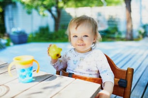 Cute baby girl eating apple at the outdoor table on a warm summer day. Little kid tasting solids at home. Baby led weaning