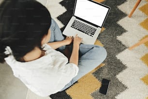 Chica hipster con estilo que usa una computadora portátil con pantalla en blanco, sentada en el piso en una habitación moderna. Maqueta de mujer joven comprando o trabajando en línea desde casa. Freelance y freelance. Vista superior