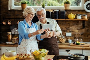 Feliz pareja madura usando tableta digital mientras prepara comida en la cocina.