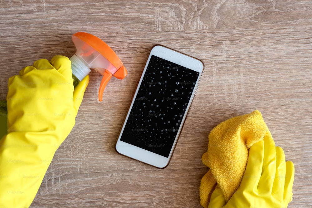 Female hand in rubber protective glove with rag wiping smartphone using cleaning spray.