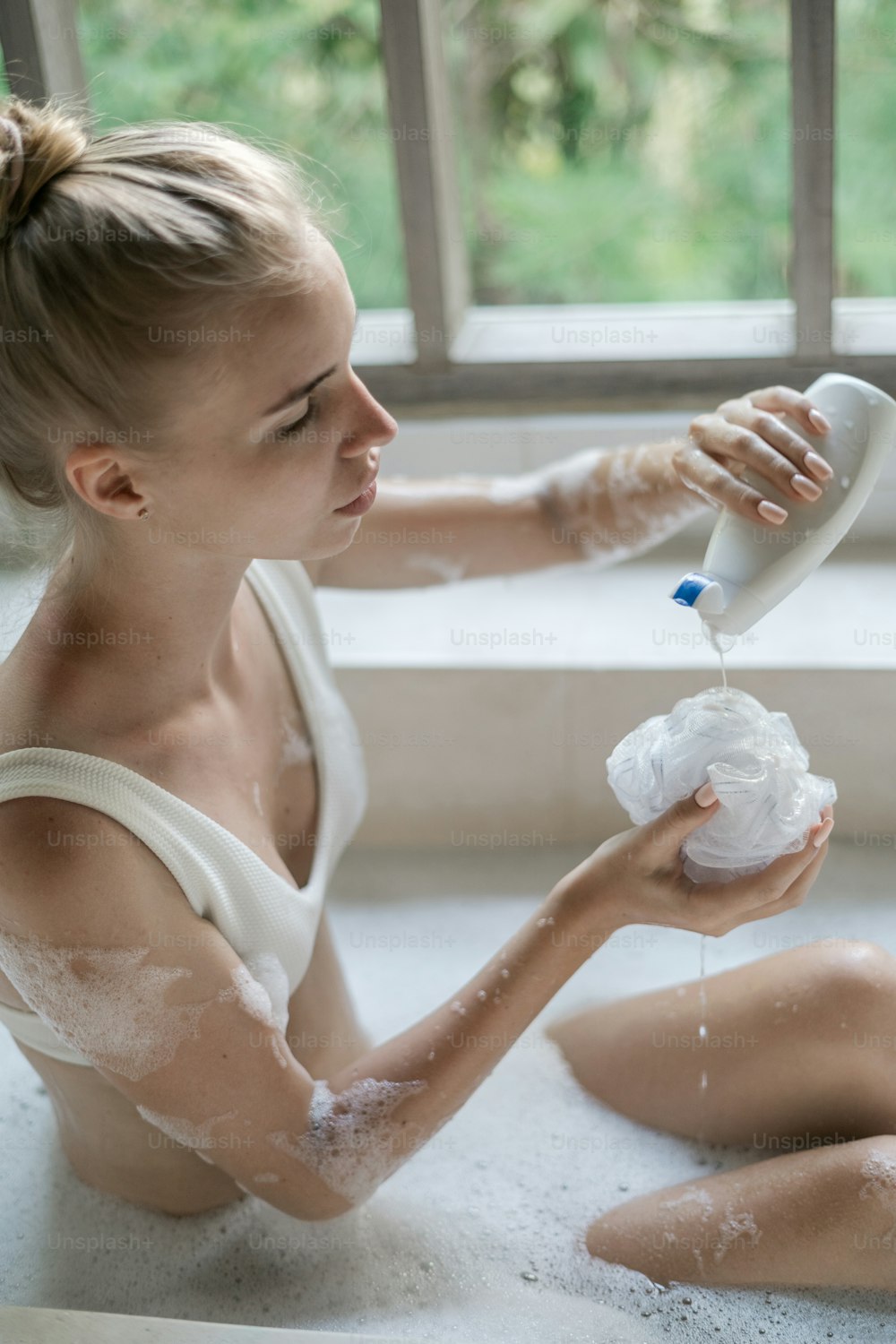 Concetto di cura della pelle. Foto verticale di giovane ragazza adulta che fa il bagno, versando gel doccia sulla spugna, sdraiata in acqua saponata a bolle, godendo della procedura di routine mattutina