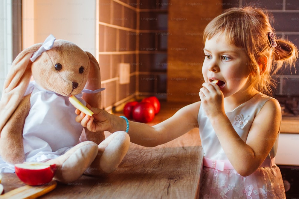 Petite fille jouant avec une peluche de lapin en le nourrissant avec une tranche de pomme fraîche dans la cuisine. Mise au point sélective.