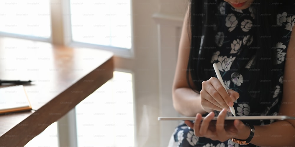 Cropped image of beautiful woman holding a computer tablet and stylus pen while sitting at the wooden working desk over comfortable living room as background.