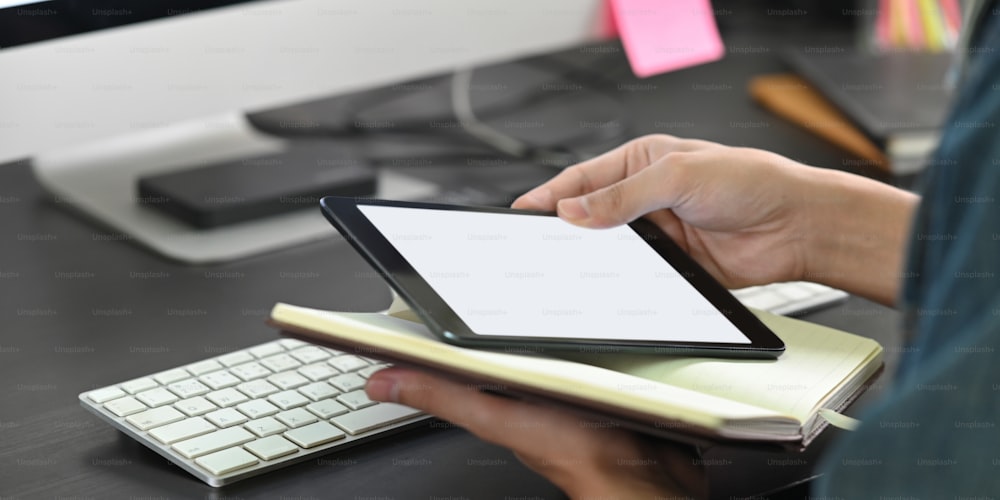 Cropped image of smart man's hands holding a white blank screen computer tablet and notebook while sitting at the working desk over wireless keyboard and computer monitor as background.